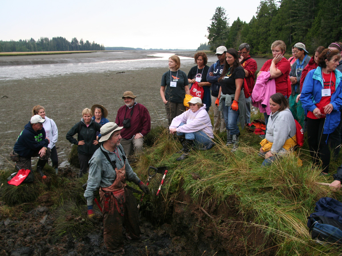 CEETEP participants learn about tsunami geology from US Geological Survey research, Brian Atwater. Niawiakum River, Washington. Participants are gathered at the edge of the salt marsh as Brian explains the layers observed below.